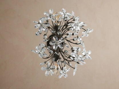 Fiore Sconce, a dark metal wall light with many clear crystal flowers & leaf accents, glowing warmly on a tan wall.