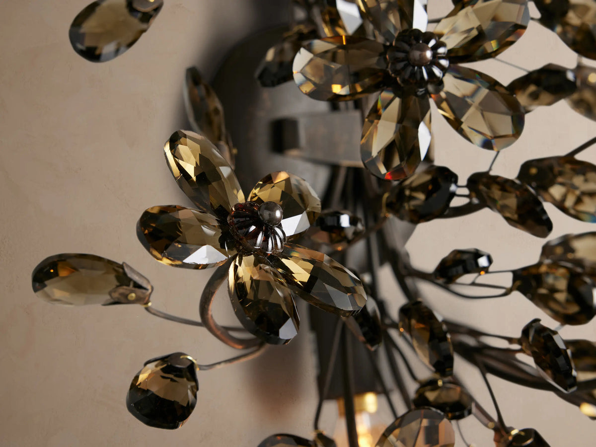Fiore Sconce: dark iron floral wall light with numerous smoky brown crystal flower petals and glowing bulbs on a beige wall.