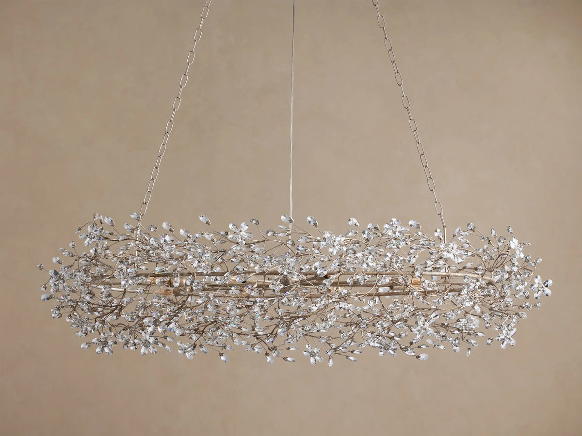Fiore Oval Chandelier, silver-toned oval metal frame intricately adorned with numerous small clear crystals, illuminated, against a neutral wall.