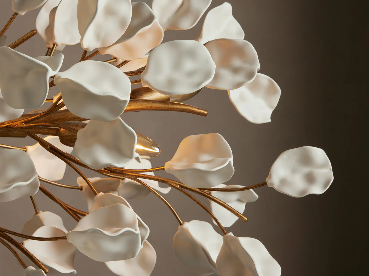 Lunaria Linear Chandelier; white leaf-shaped elements on gold branches, suspended by two gold chains against a beige wall.