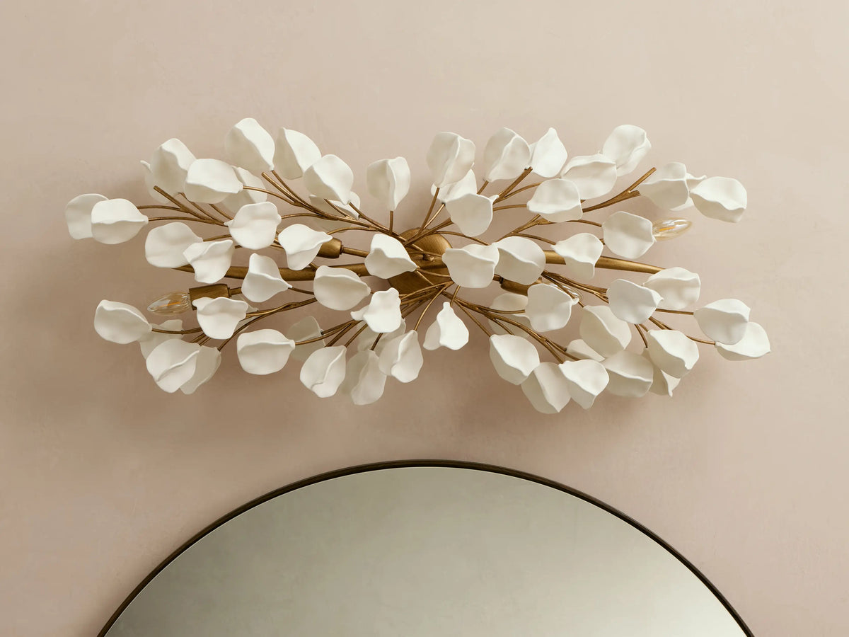 Lunaria Grand Sconce: Horizontal, white petal-like leaves on golden branches, glowing warm lights, above a round mirror.