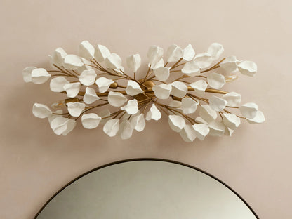 Lunaria Grand Sconce: Horizontal, white petal-like leaves on golden branches, glowing warm lights, above a round mirror.