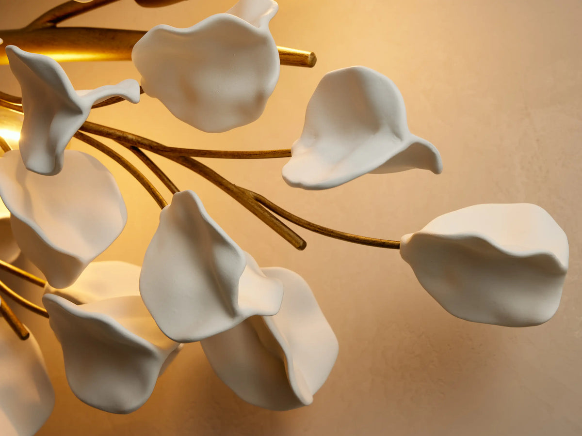 Lunaria Grand Sconce: A horizontal gold sconce with numerous white ceramic leaf-like forms and visible bulbs, on a light-pink wall.