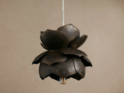 Lorca Pendant, a layered dark bronze metal flower light with distressed brass accents and a warm glowing bulb against a brown wall.