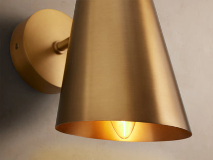 Elih Sconce, a modern wall-mounted light with a brushed brass finish, features a round backplate and a conical shade.