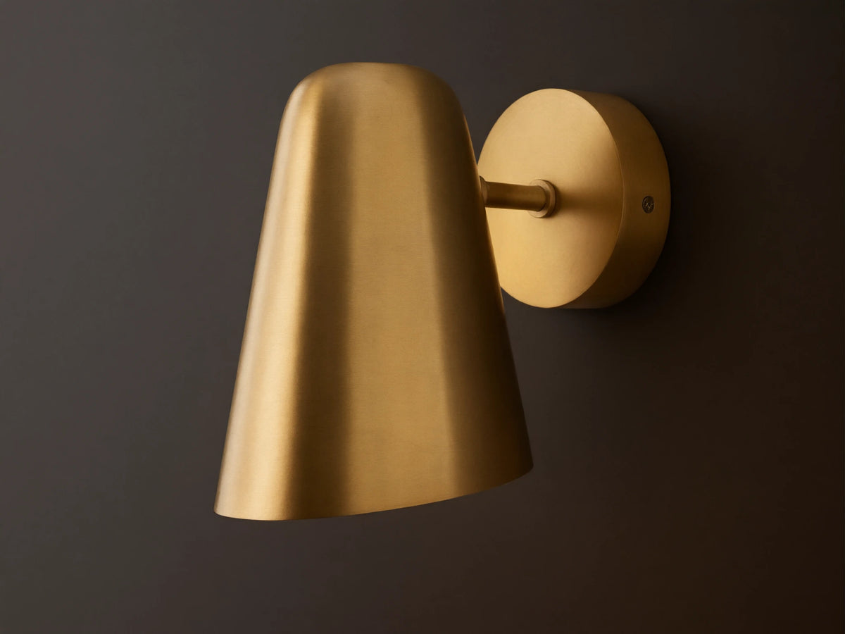 Elih Sconce, a brushed brass wall-mounted light fixture with a circular backplate and a downward-facing conical shade on a textured wall.
