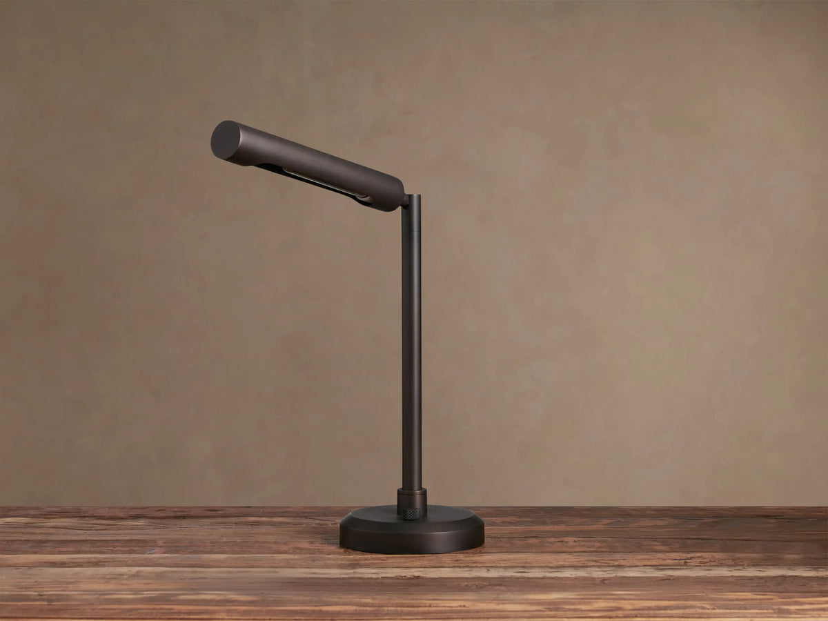 Elih Desk Lamp: Antiqued bronze brass finish, modern design with cylindrical head angled down, emitting warm light on a wooden surface.