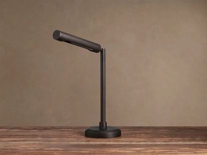 Elih Desk Lamp: Antiqued bronze brass finish, modern design with cylindrical head angled down, emitting warm light on a wooden surface.