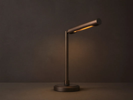 Elih Desk Lamp: Antiqued bronze brass finish, modern design with cylindrical head angled down, emitting warm light on a wooden surface.
