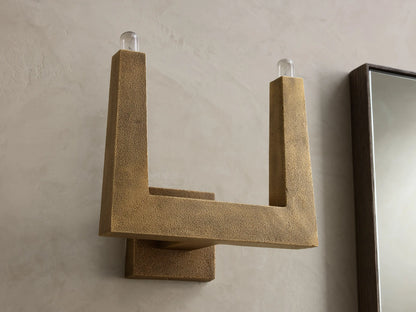 Golden textured wall sconce with an L-shaped arm and a lit bulb at the top, casting warm light on the wall.
