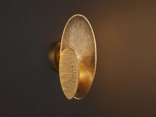 Udaya Embossed Sconce, golden, with overlapping oval plates; one radial, one horizontal embossed. Warm light on a wall.