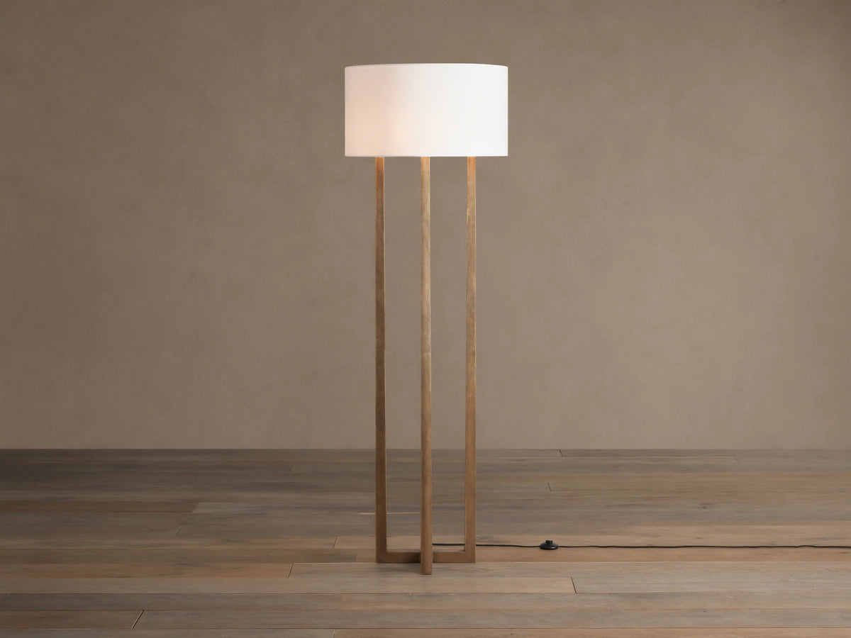 Emmet Floor Lamp with a white linen drum shade & three antiqued brass/bronze vertical columns, standing on a wood floor.