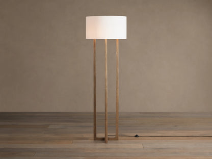 Emmet Floor Lamp with a white linen drum shade & three antiqued brass/bronze vertical columns, standing on a wood floor.