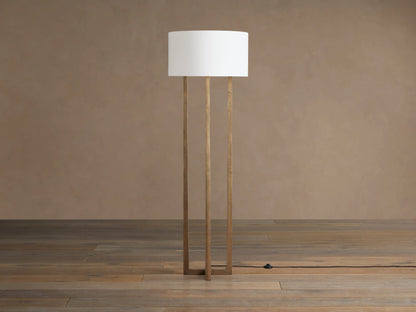 Emmet Floor Lamp with three antiqued brass rectangular legs and white linen drum shade, on wooden floor against a brown wall.