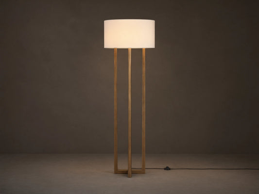 Emmet Floor Lamp with a white linen drum shade & three antiqued brass/bronze vertical columns, standing on a wood floor.