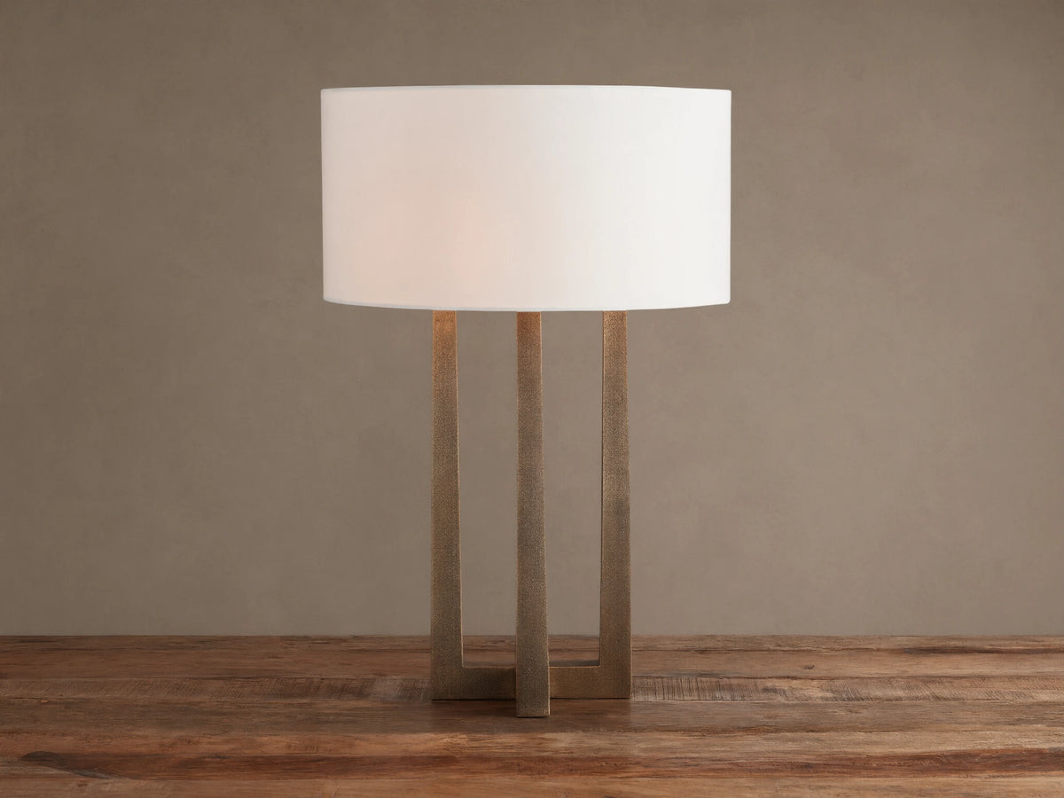 Emmet Table Lamp: lit white cylindrical linen shade, textured antiqued brass three-legged base, on a rustic wood table.