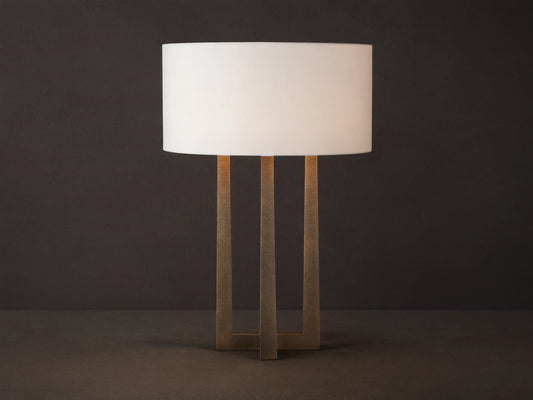 Emmet Table Lamp: lit white cylindrical linen shade, textured antiqued brass three-legged base, on a rustic wood table.
