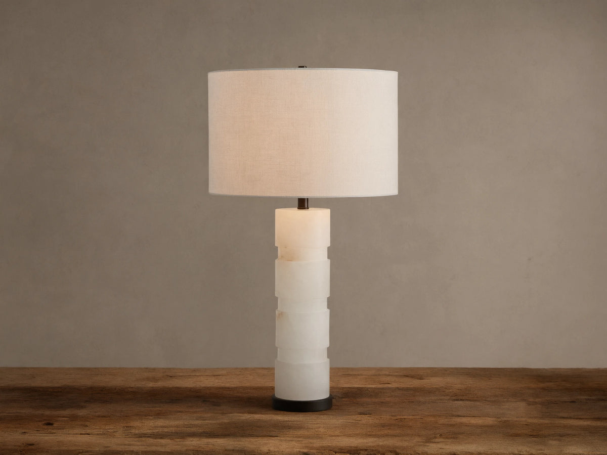 Novalie Table Lamp: Carved white alabaster body with bronze base, topped by a large off-white fabric drum shade on a wooden surface.