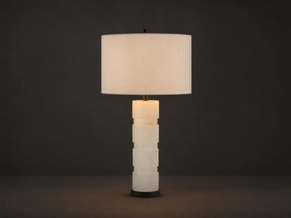 Novalie Table Lamp: Carved white alabaster body with bronze base, topped by a large off-white fabric drum shade on a wooden surface.
