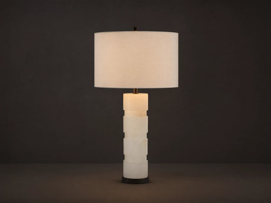 Novalie Table Lamp: Carved white alabaster body with bronze base, topped by a large off-white fabric drum shade on a wooden surface.