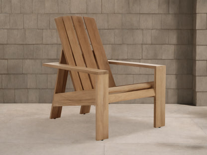 Natural teak wood Adirondack Outdoor Lounge Chair, angled slightly right, with a slatted back and wide arms, against a wooden shingle wall.