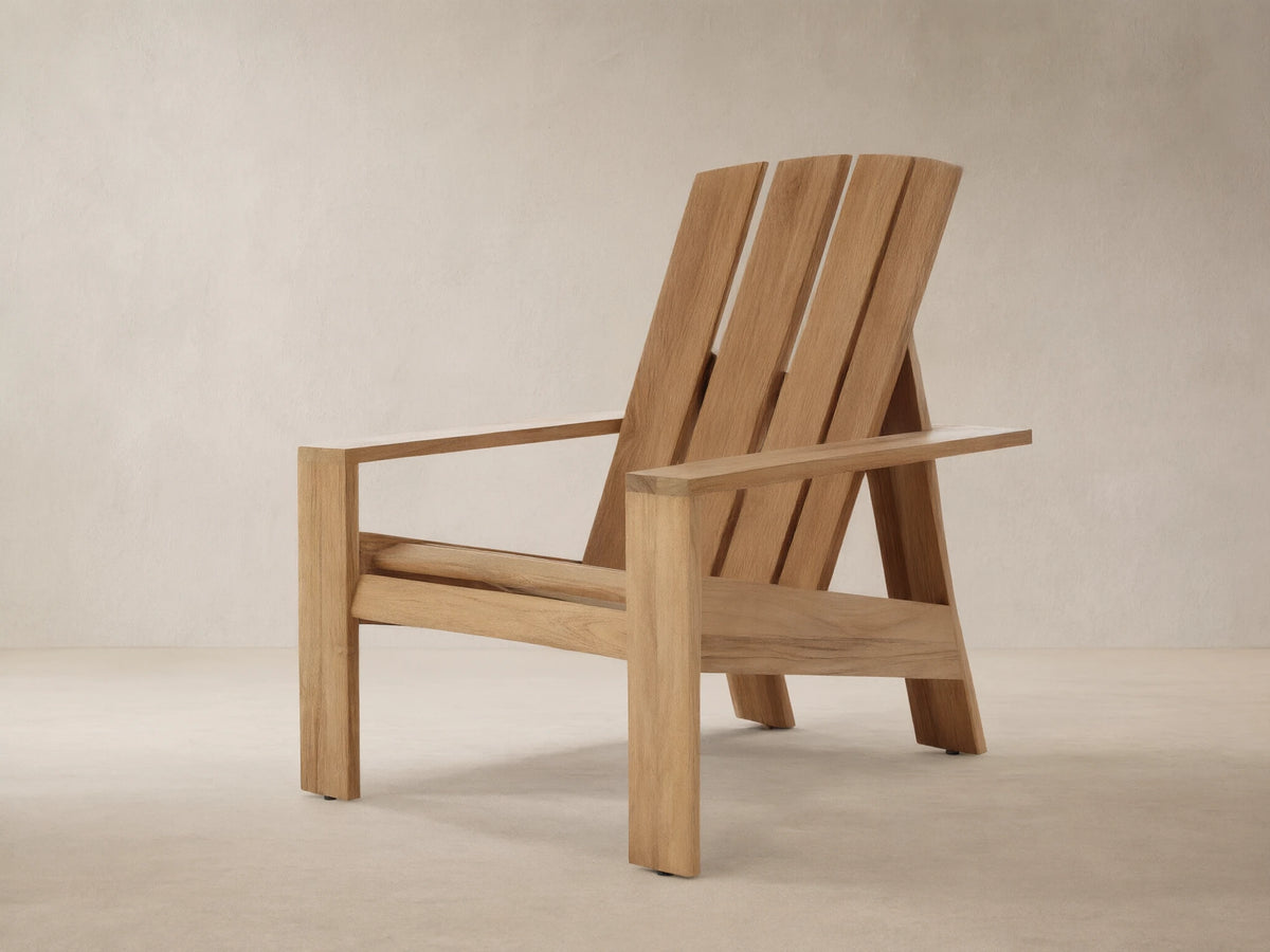 Natural teak wood Adirondack Outdoor Lounge Chair, angled slightly right, with a slatted back and wide arms, against a wooden shingle wall.