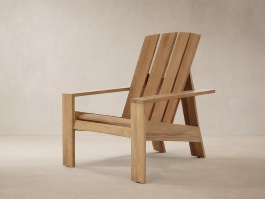 Natural teak wood Adirondack Outdoor Lounge Chair, angled slightly right, with a slatted back and wide arms, against a wooden shingle wall.