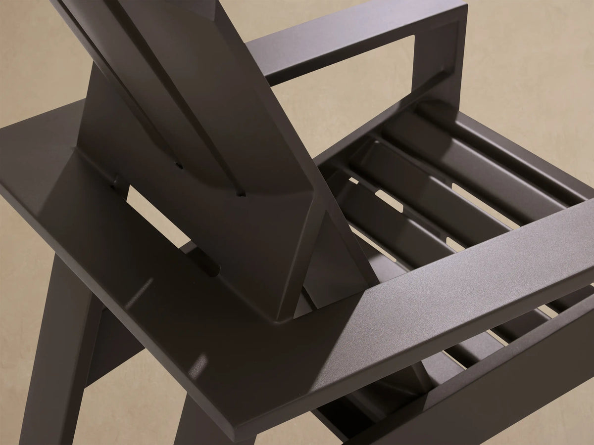 Front view of a dark gray aluminum Adirondack lounge chair with slatted back & wide arms, on a patio against a cedar wall.