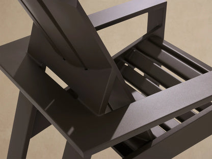 Front view of a dark gray aluminum Adirondack lounge chair with slatted back & wide arms, on a patio against a cedar wall.