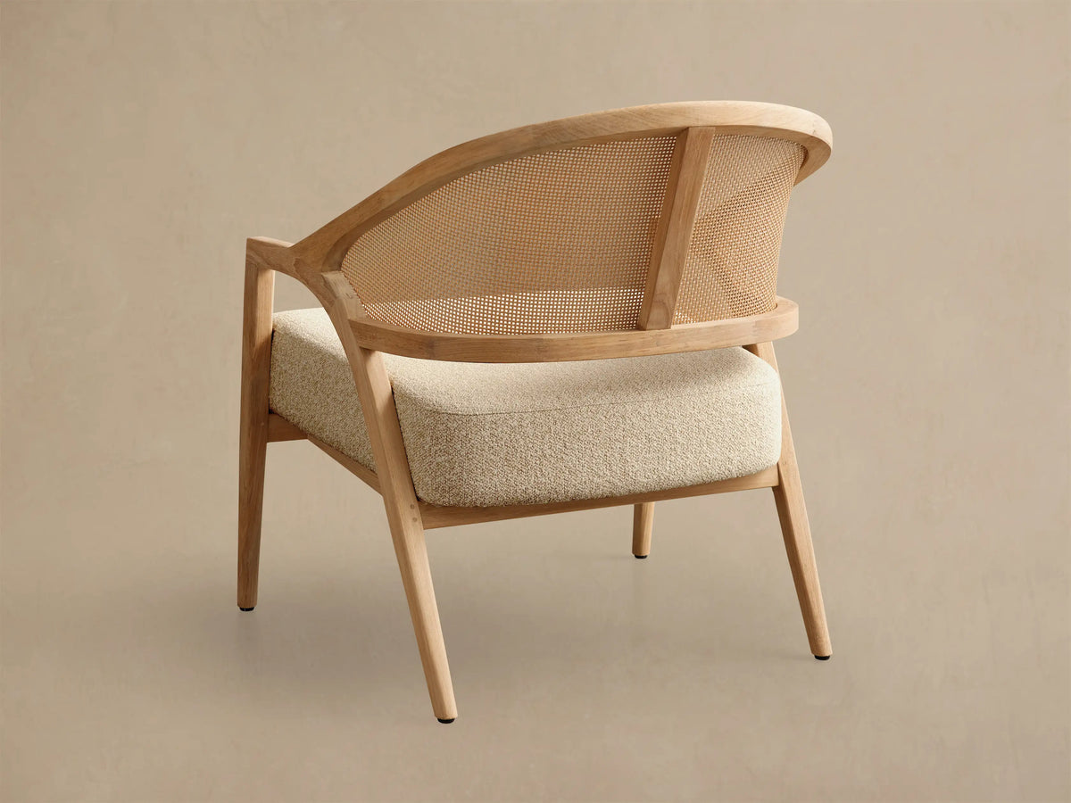 Close-up of the natural teak Aimee Outdoor Lounge Chair, angled view showing curved frame, woven resin back, and light beige cushion.