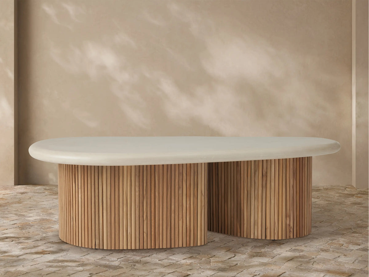 Alesso Outdoor Coffee Table with oval off-white top, two semicircular bases of vertical natural teak slats on paved ground.