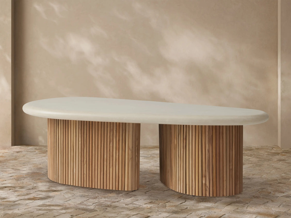 Alesso Outdoor Coffee Table with oval off-white top, two semicircular bases of vertical natural teak slats on paved ground.