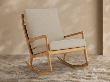 Altea Outdoor Rocking Chair with light teak wood frame and light beige cushions, angled on a stone tile floor.