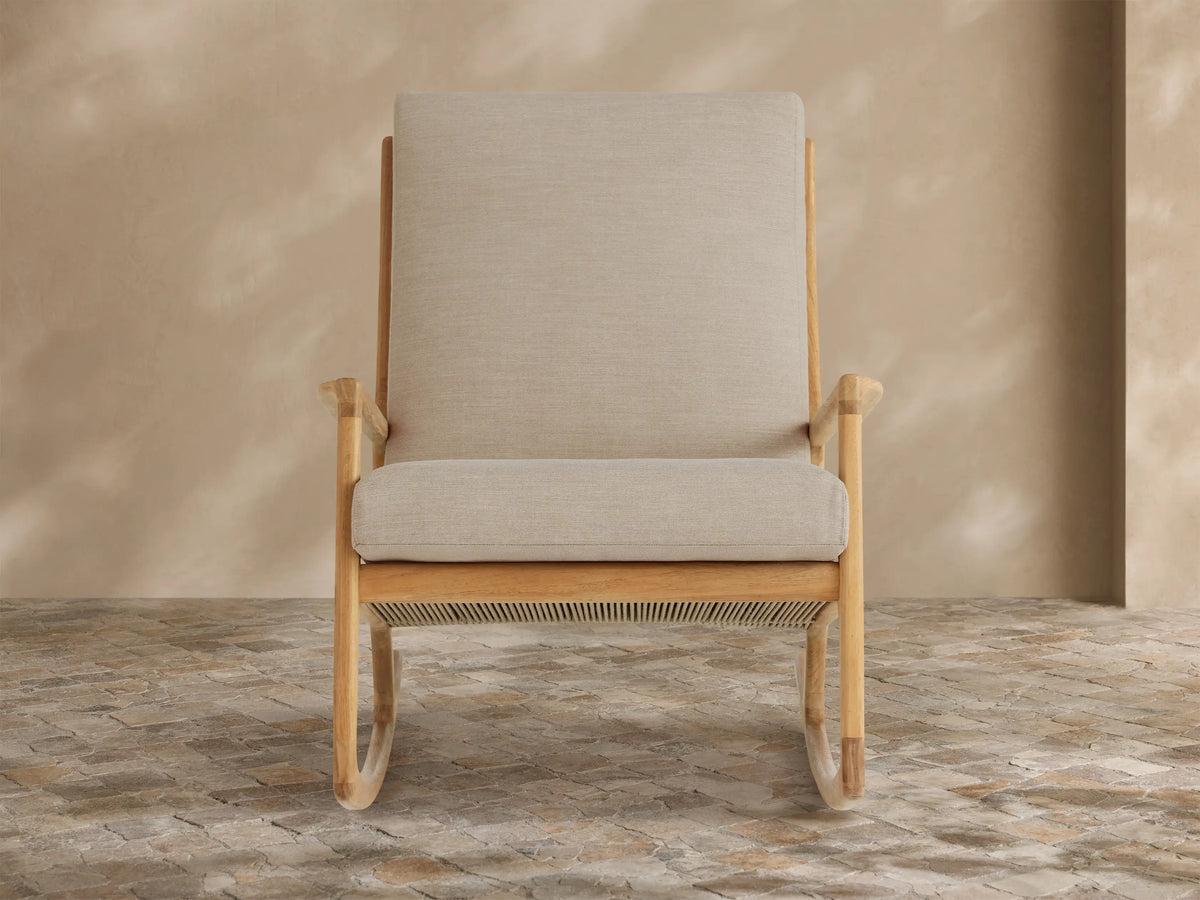 Altea Outdoor Rocking Chair with light teak wood frame and light beige cushions, angled on a stone tile floor.