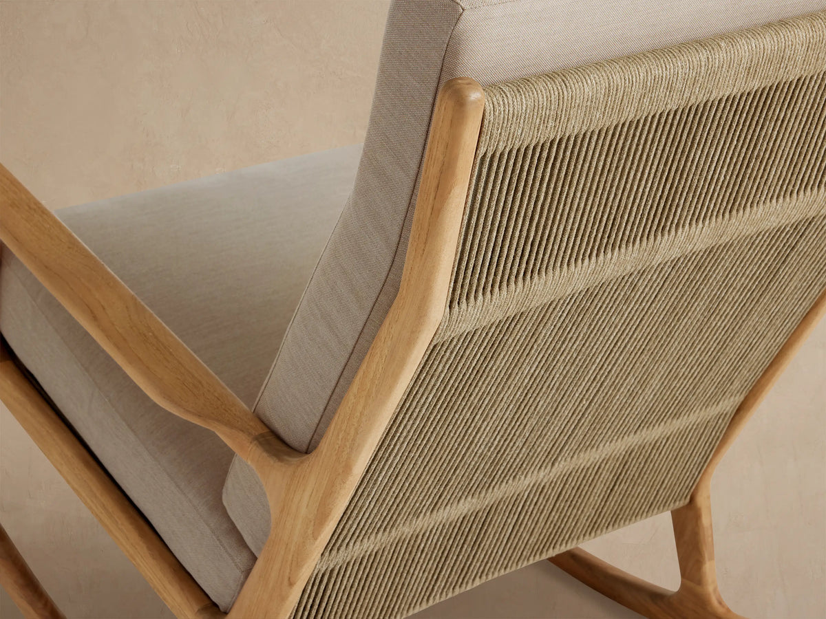 Front view of an Altea Outdoor Rocking Chair with a light teak frame, beige cushions, and woven rope, on a stone floor.