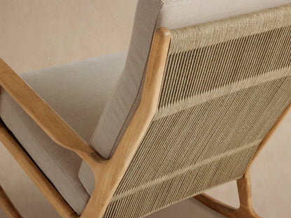 Front view of an Altea Outdoor Rocking Chair with a light teak frame, beige cushions, and woven rope, on a stone floor.
