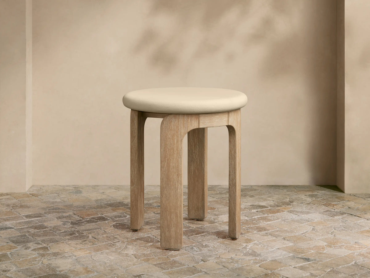 Anami Outdoor End Table with a light, round top and four curved, natural teak wood legs on a tiled floor.