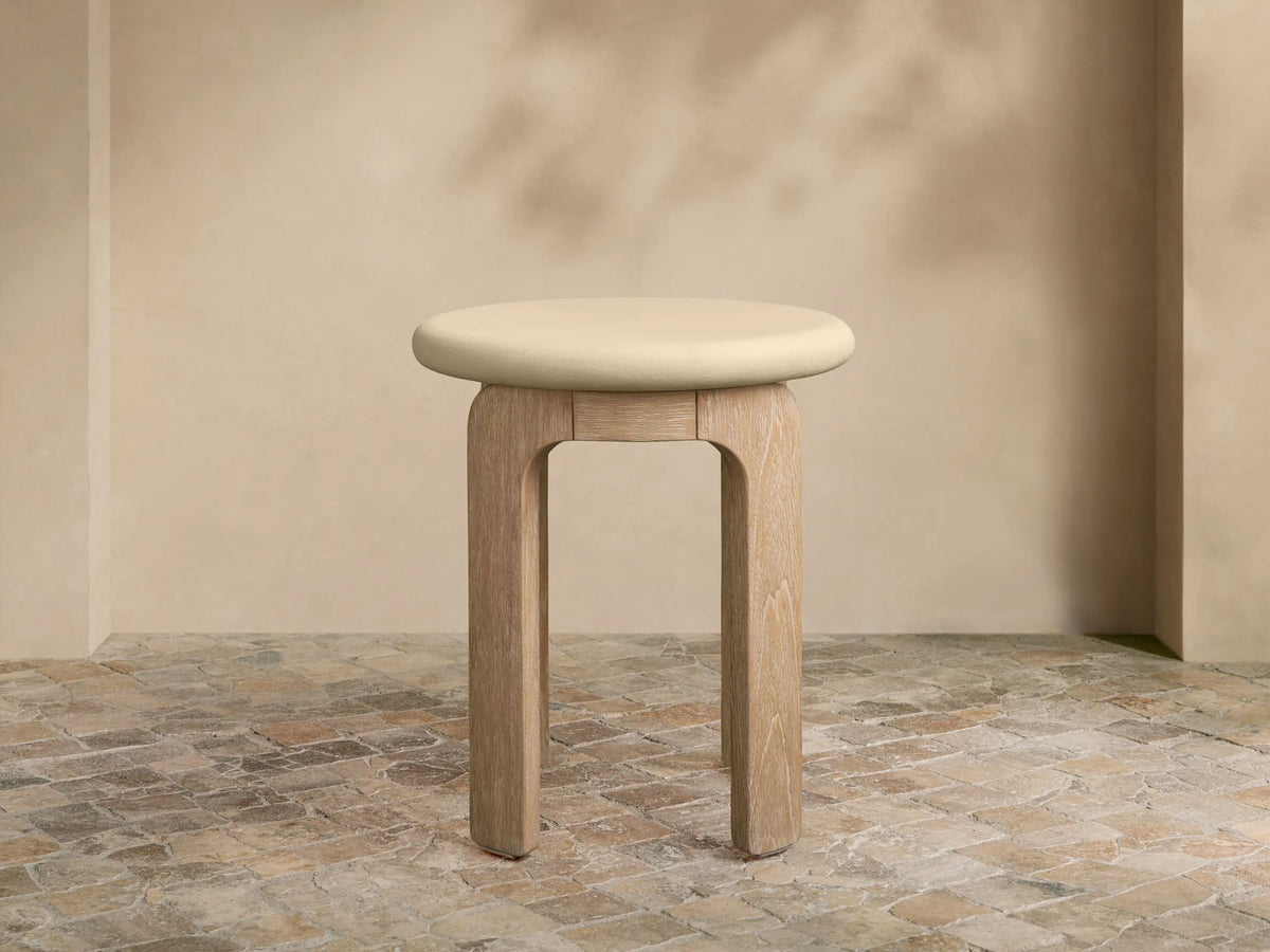 Anami Outdoor End Table with a light, round top and four curved, natural teak wood legs on a tiled floor.