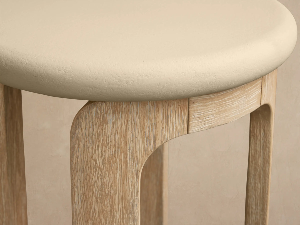 Anami Outdoor End Table: round cream top, light natural teak U-shaped legs on a stone floor, against a beige wall.