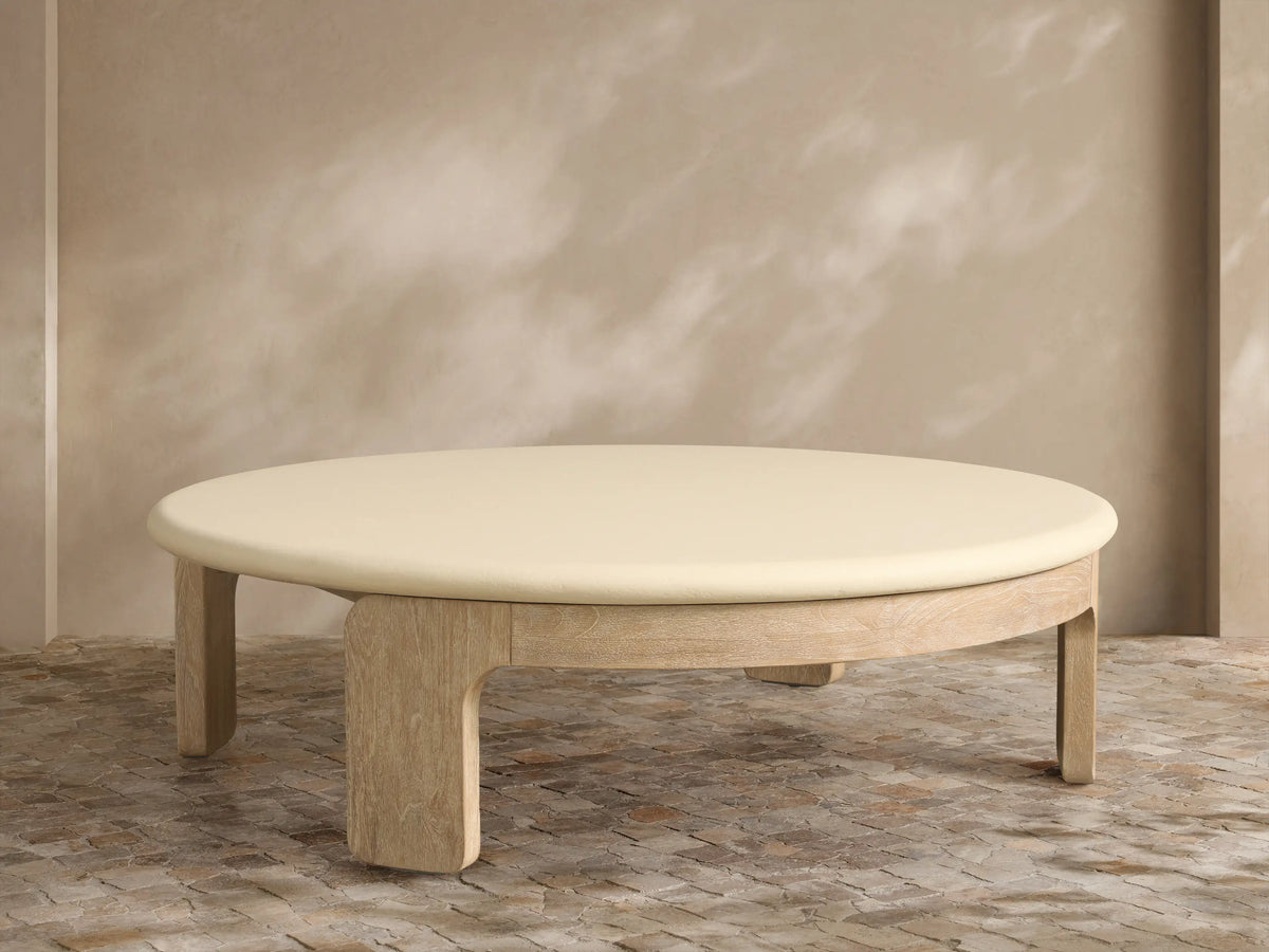 Anami Outdoor Round Coffee Table with a light wood base and an off-white round top, on a stone patio against a textured wall.