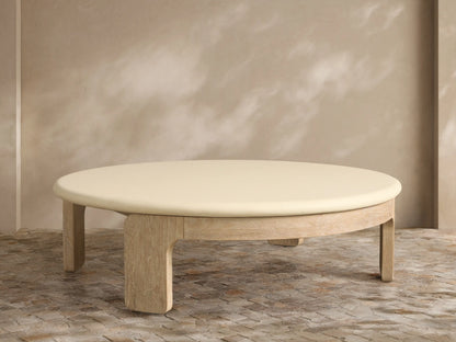 Anami Outdoor Round Coffee Table with a light wood base and an off-white round top, on a stone patio against a textured wall.