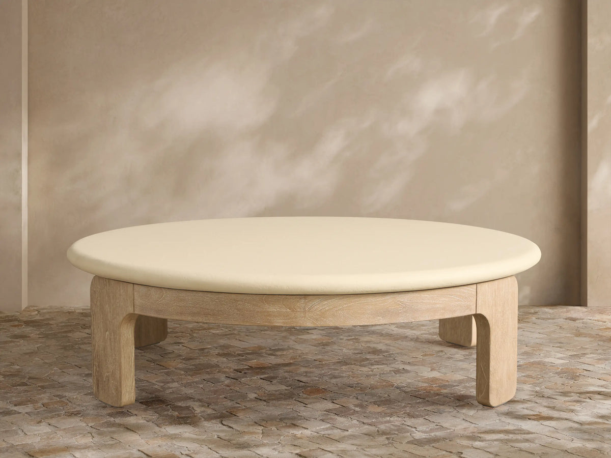 Anami Outdoor Round Coffee Table with a light wood base and an off-white round top, on a stone patio against a textured wall.