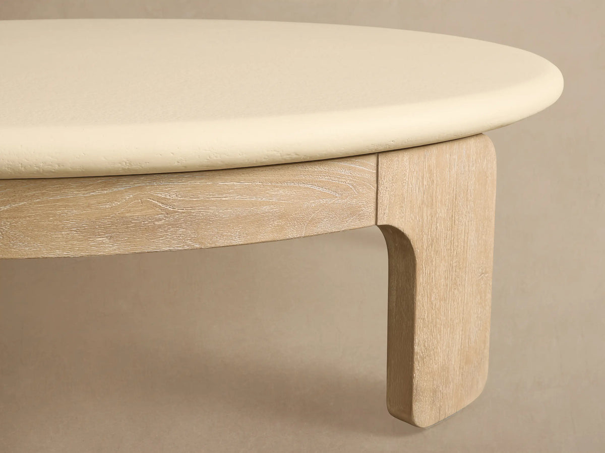 Anami Outdoor Round Coffee Table with a light natural wood base and a smooth cream-colored round top on a tiled patio.