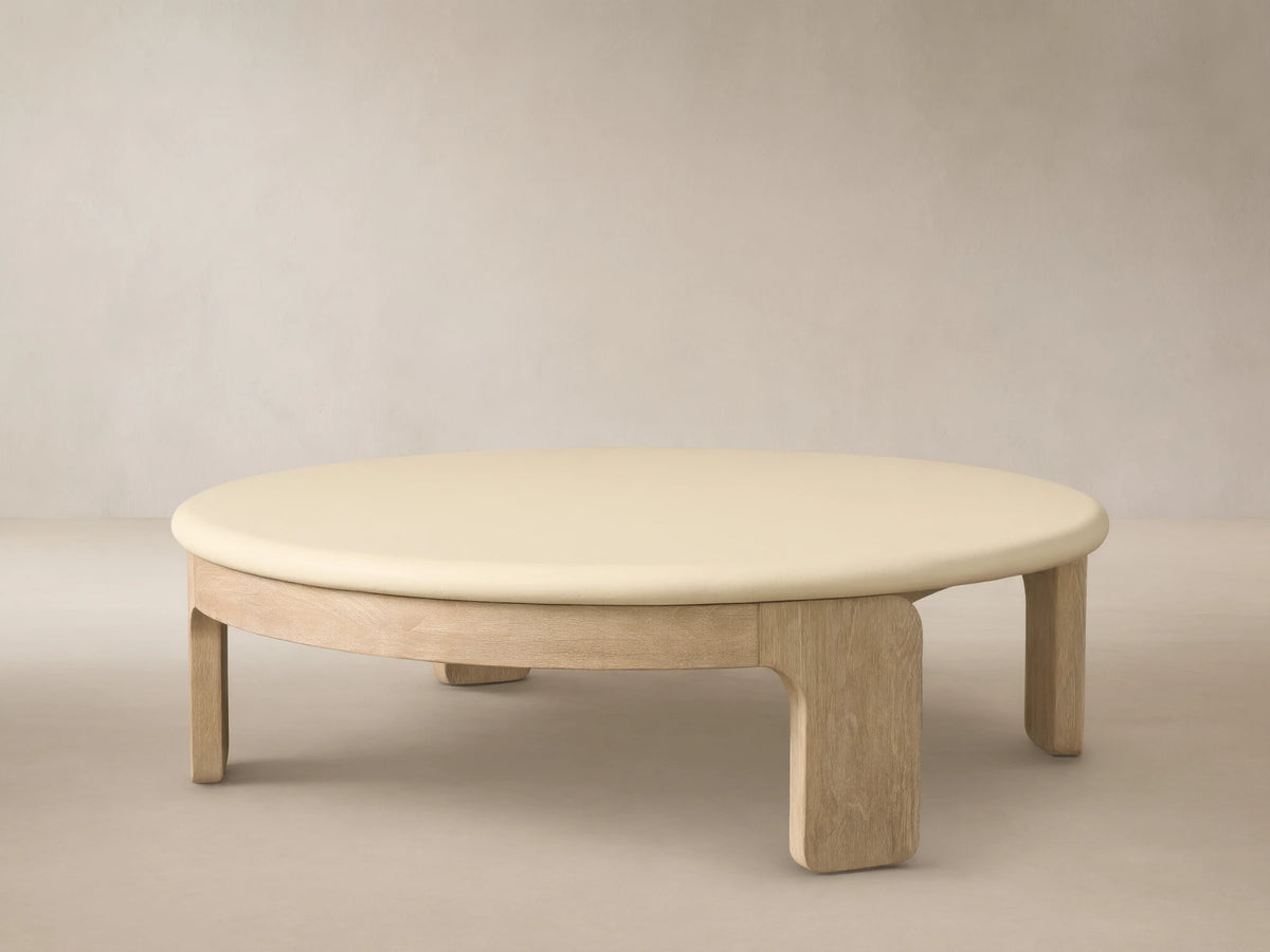 Anami Outdoor Round Coffee Table with a light wood base and an off-white round top, on a stone patio against a textured wall.