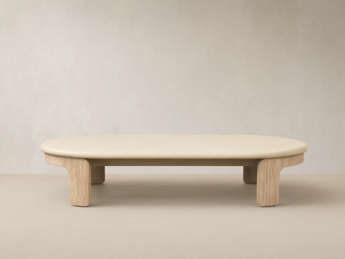 Anami Outdoor Coffee Table with an oval, cream-colored top on natural teak wood legs, on a tiled patio floor.