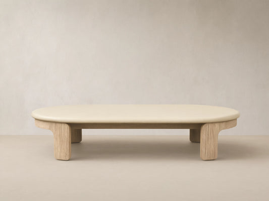 Anami Outdoor Coffee Table with an oval, cream-colored top on natural teak wood legs, on a tiled patio floor.
