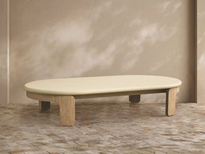 Anami Outdoor Coffee Table with an oval, cream-colored top on natural teak wood legs, on a tiled patio floor.