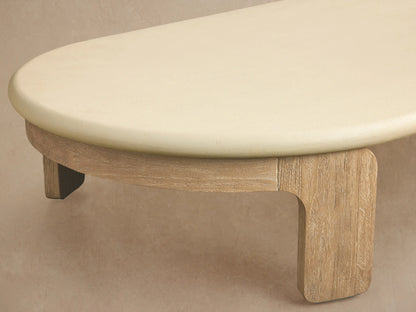 Anami Outdoor Coffee Table: low, oval cream top on light natural teak wood slab legs, on a textured stone patio.