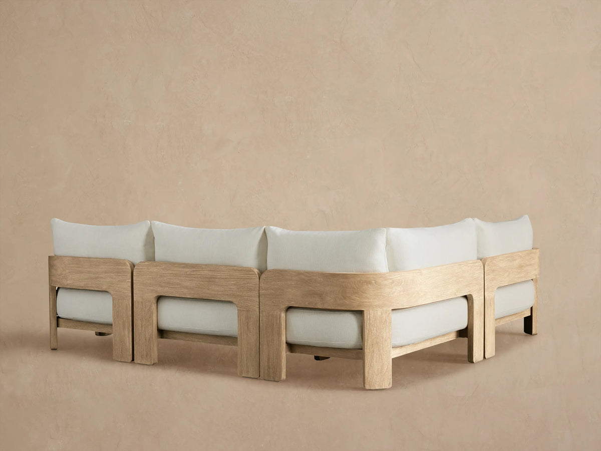 Close-up of Anami Outdoor Two-Piece Sectional armrest & cushion. Light teak wood frame with curved grain, pale grey fabric.