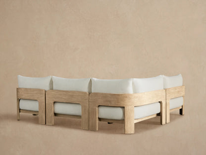 Close-up of Anami Outdoor Two-Piece Sectional armrest & cushion. Light teak wood frame with curved grain, pale grey fabric.